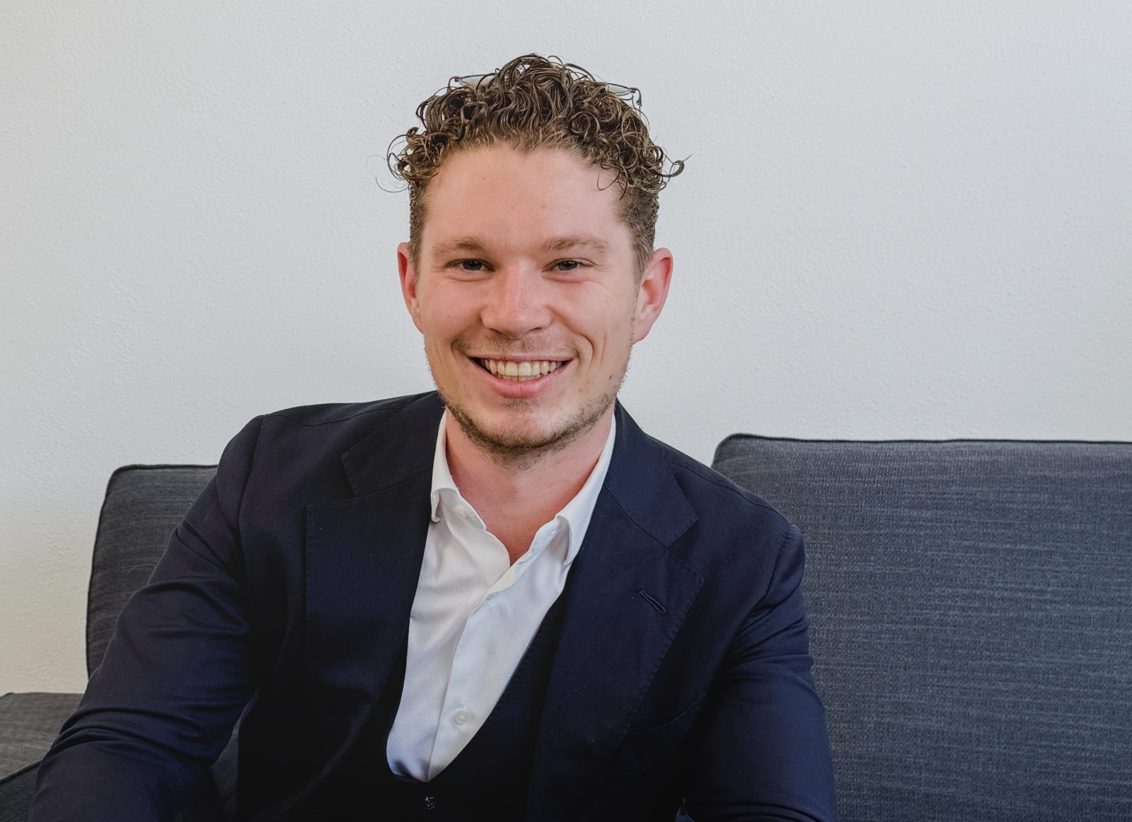 Ein junger, blonder Mann in Anzug, der Dominik Seucan ist, sitz auf einem Sofa und lächelt Richtung Kamera